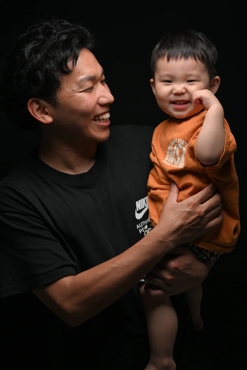 黒背景でオレンジ色の服の子どもを抱き、笑顔を向ける父親